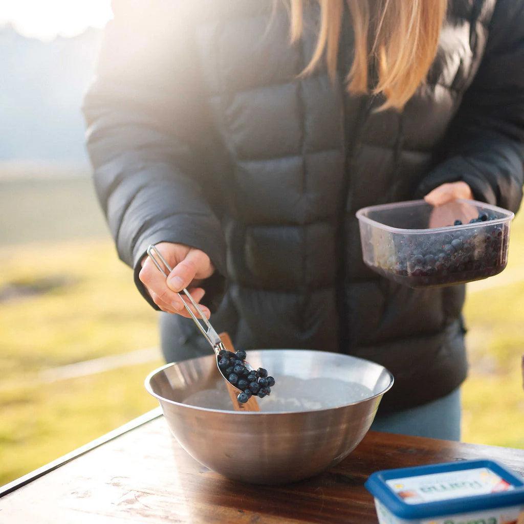 Person adding blueberries to a metal bowl with camp kitchen kit outdoors in sunlight