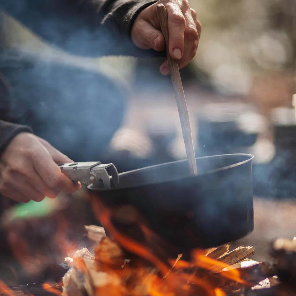 Person cooking with a black camping pot over a campfire outdoors
