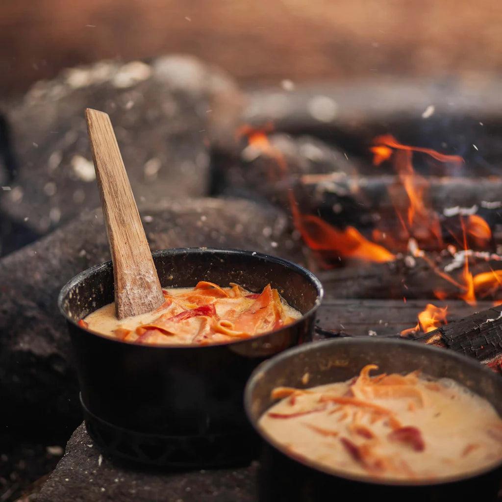 Campfire cooking with Primus pots, creamy pasta and wooden spoon at outdoor campsite.