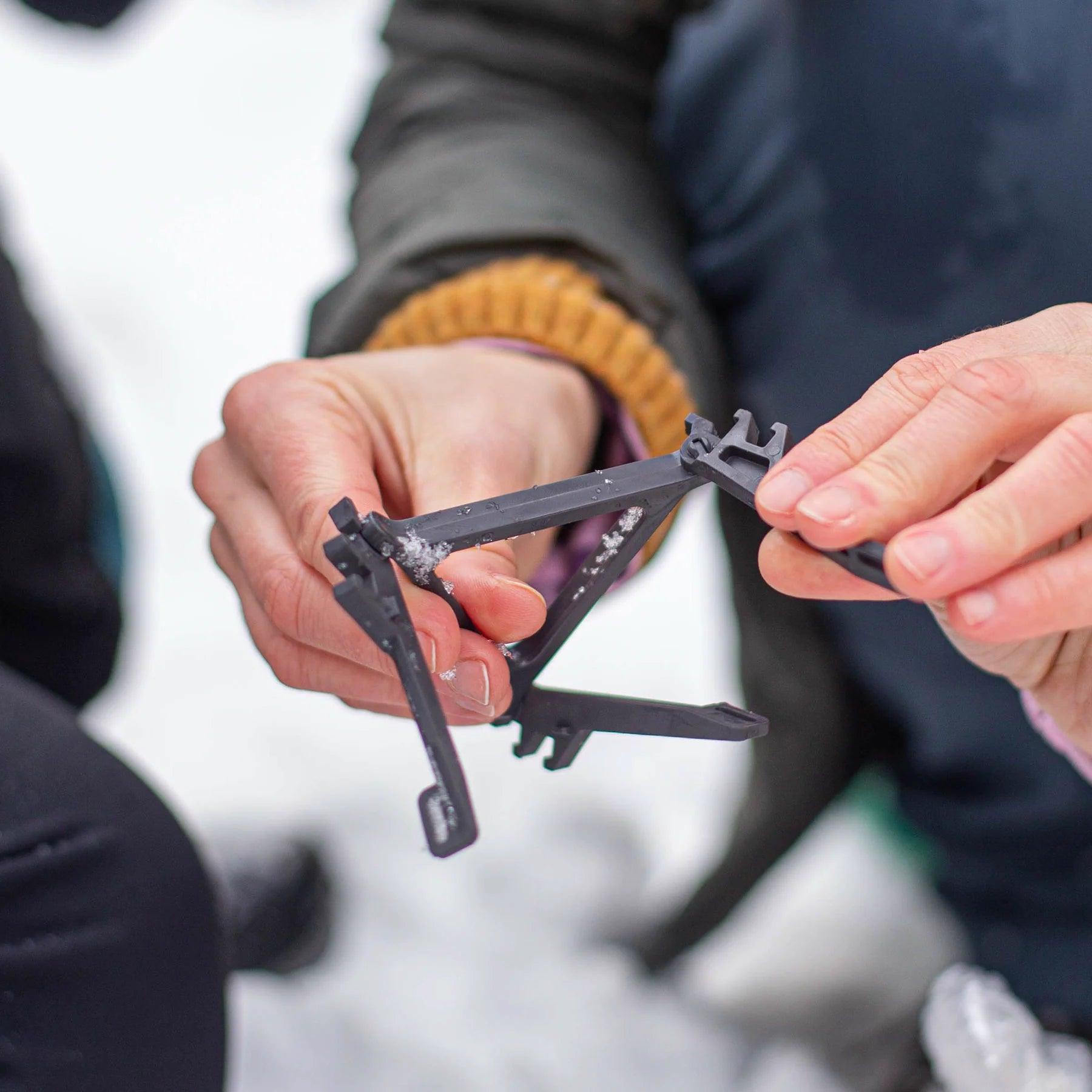 Hands assembling a Primus canister stand outdoors on snow, camping gear detail