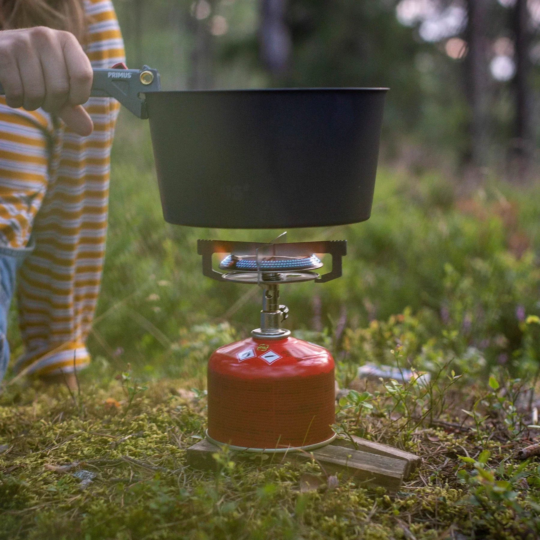 Person using Primus camping stove with black pot outdoors on forest ground