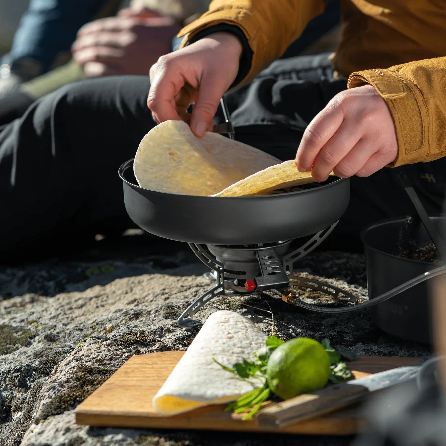 Person cooking tortillas on a portable camping stove outdoors with cutting board nearby