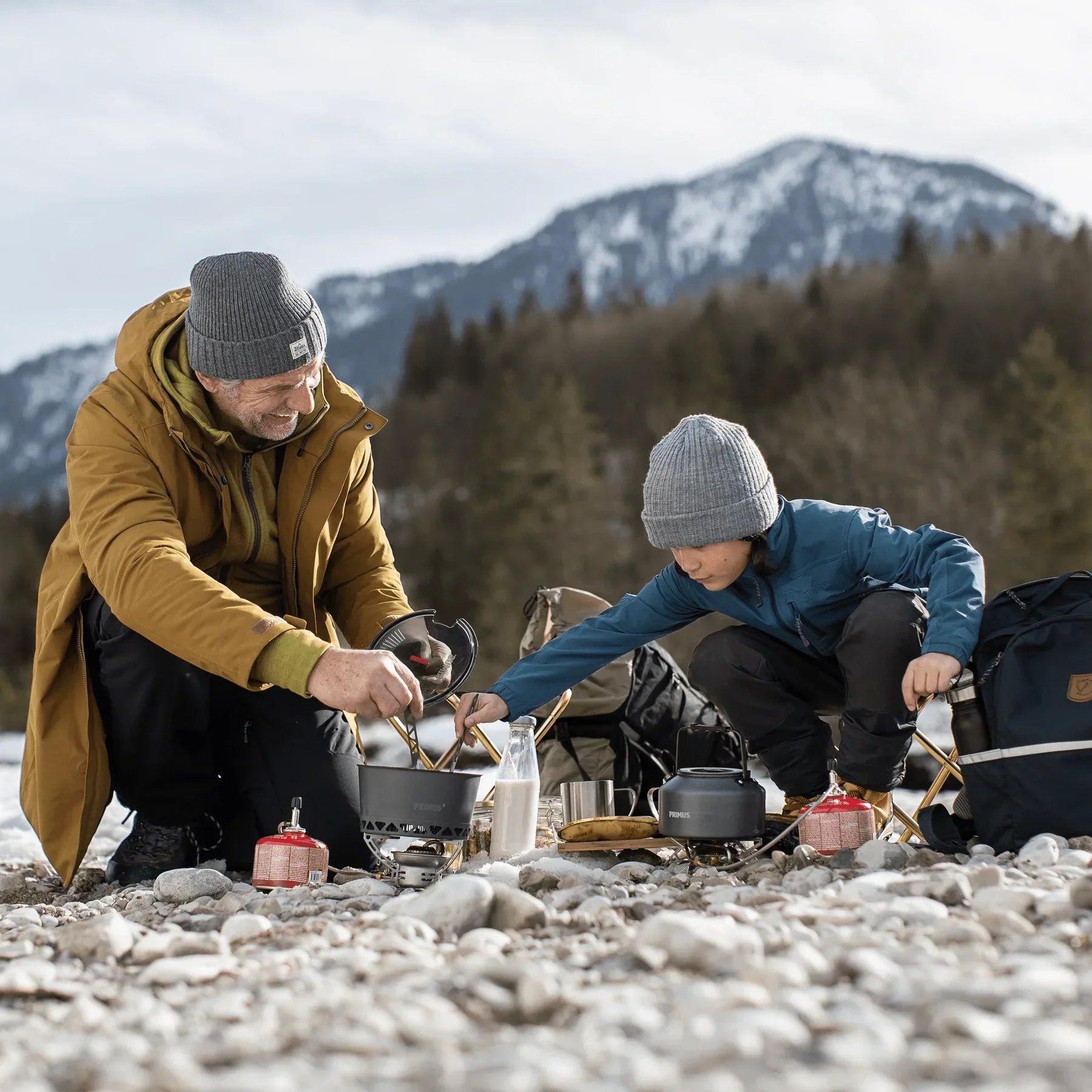 Father and son camping, using portable stove on rocky riverbank with snowy mountains