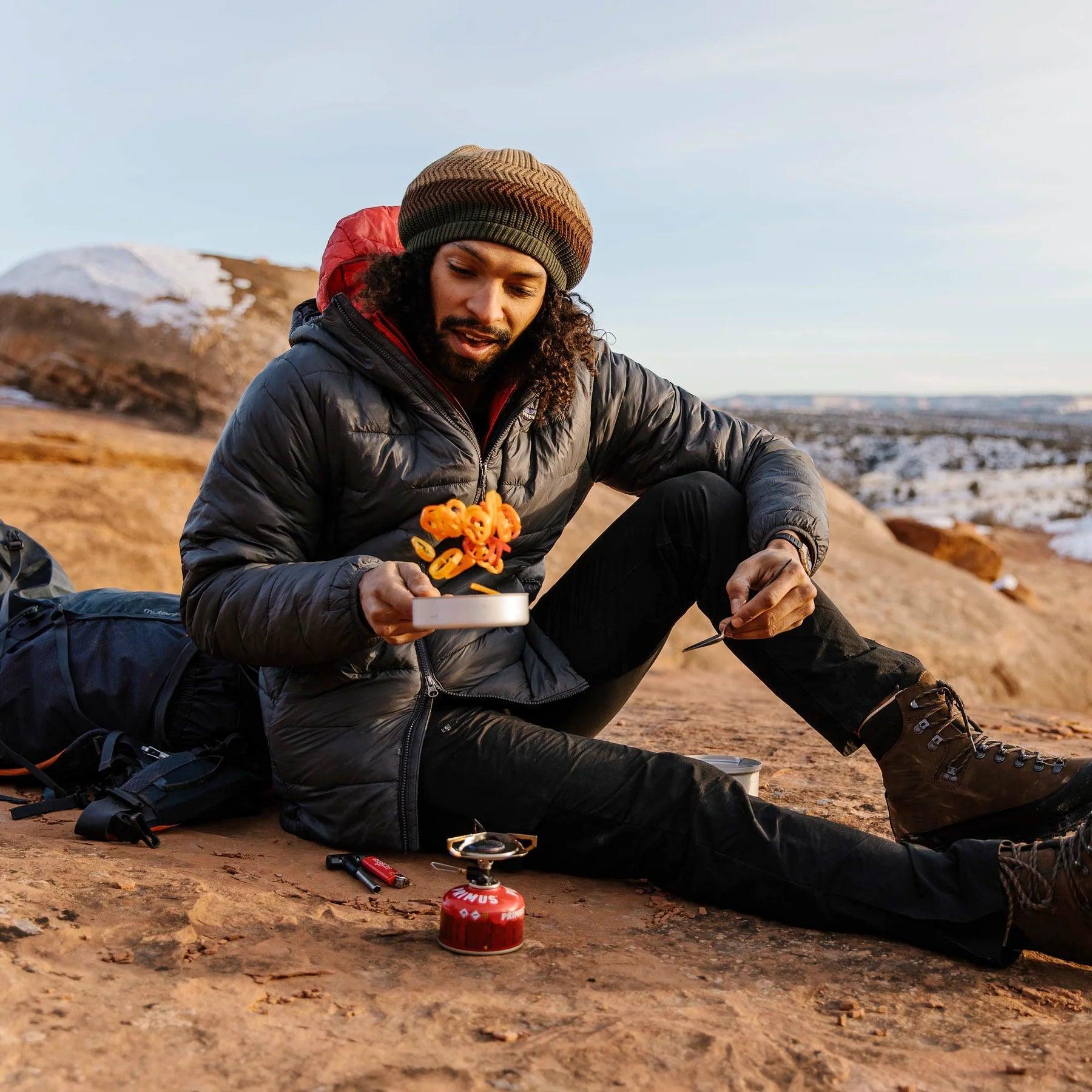 Man cooking with Primus Essential Trail Stove outdoors on rocky terrain, wearing hiking gear.
