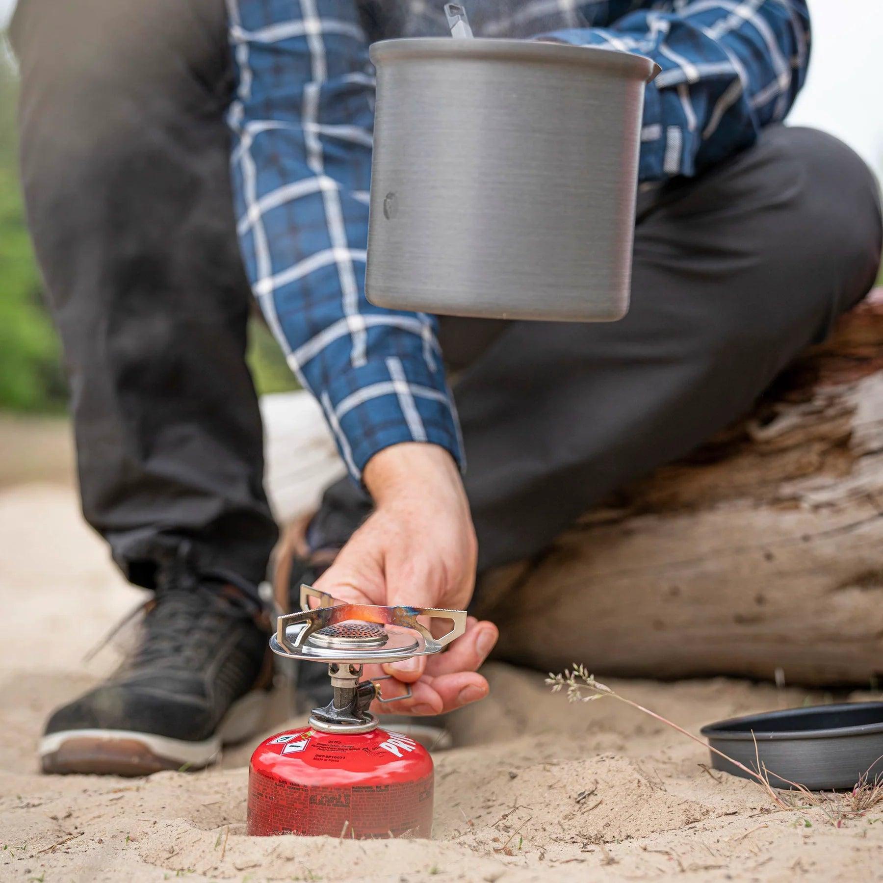 Person lighting Primus Essential Trail Stove outdoors with camping pot and log in background