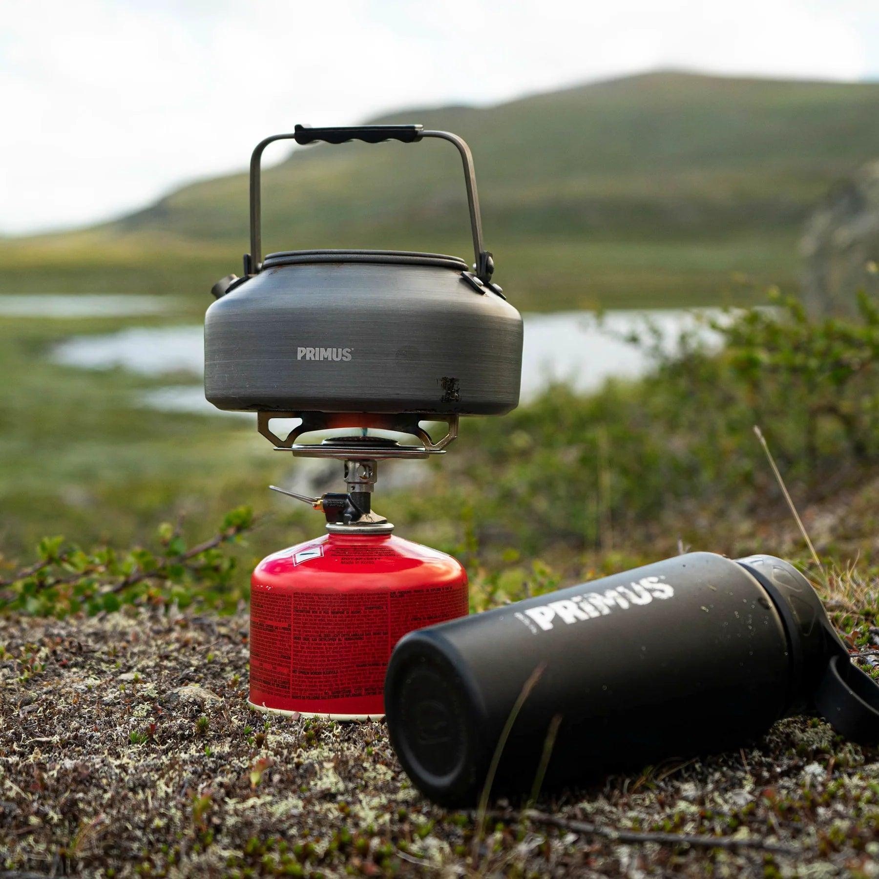 Primus Essential Trail Stove with kettle and bottle outdoors on grassy terrain near water