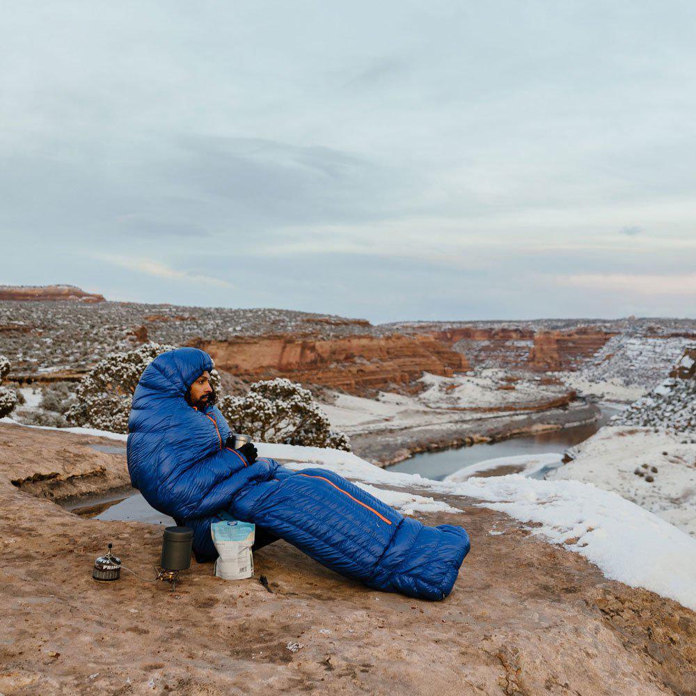 Man in blue sleeping bag camping on snowy canyon rim with winter outdoor gear.