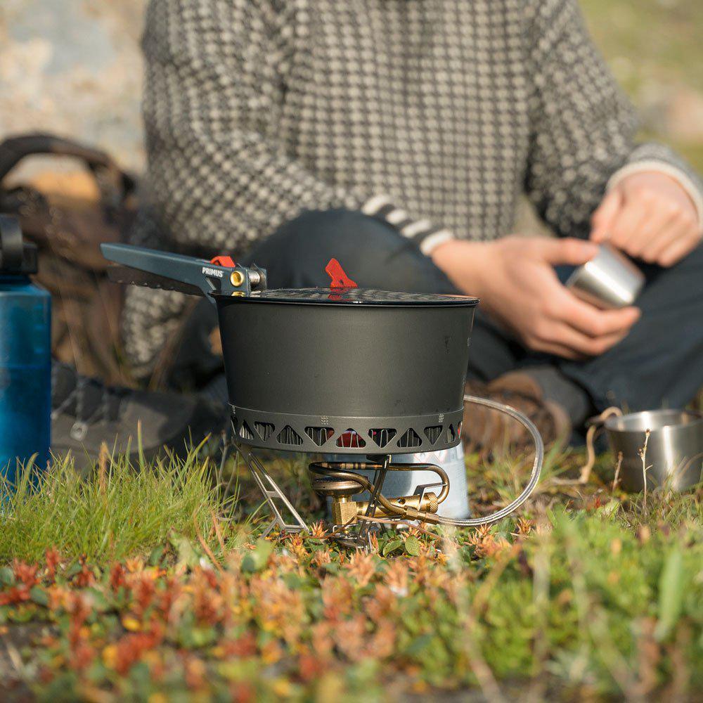 Person using Primus camping stove outdoors on grass with mug and water bottle
