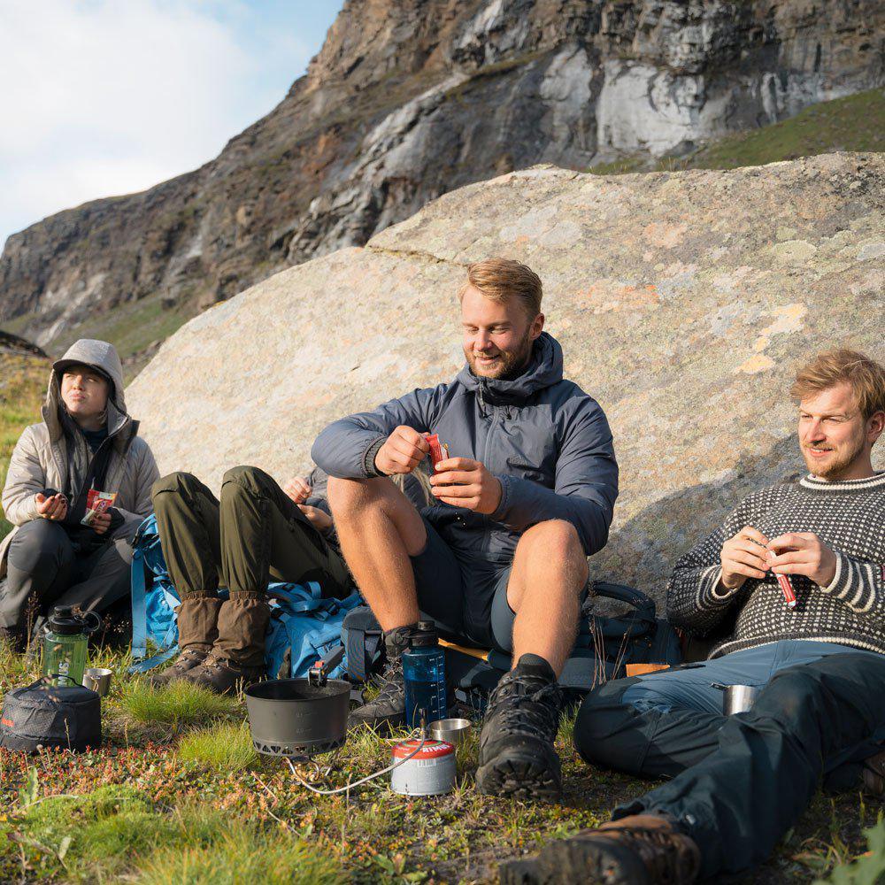 Three hikers relax by a rocky mountain, enjoying a meal with camping gear outdoors.