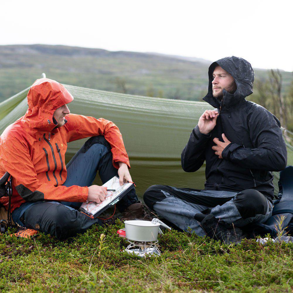 Two campers in rain jackets sit by a portable stove under a tarp on a grassy hillside.