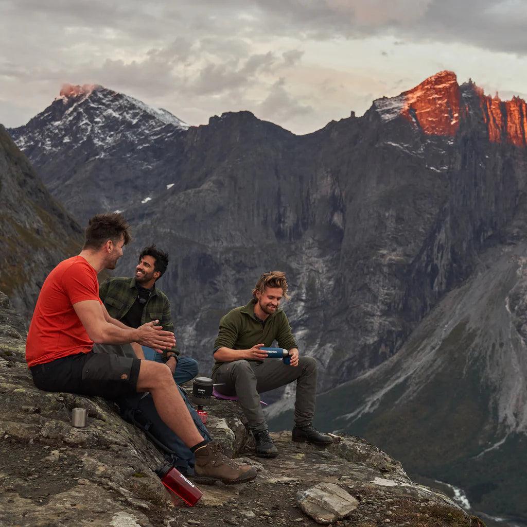 Three men camping on a rocky mountain ledge with Primus Lite XL stove, scenic alpine sunset view.