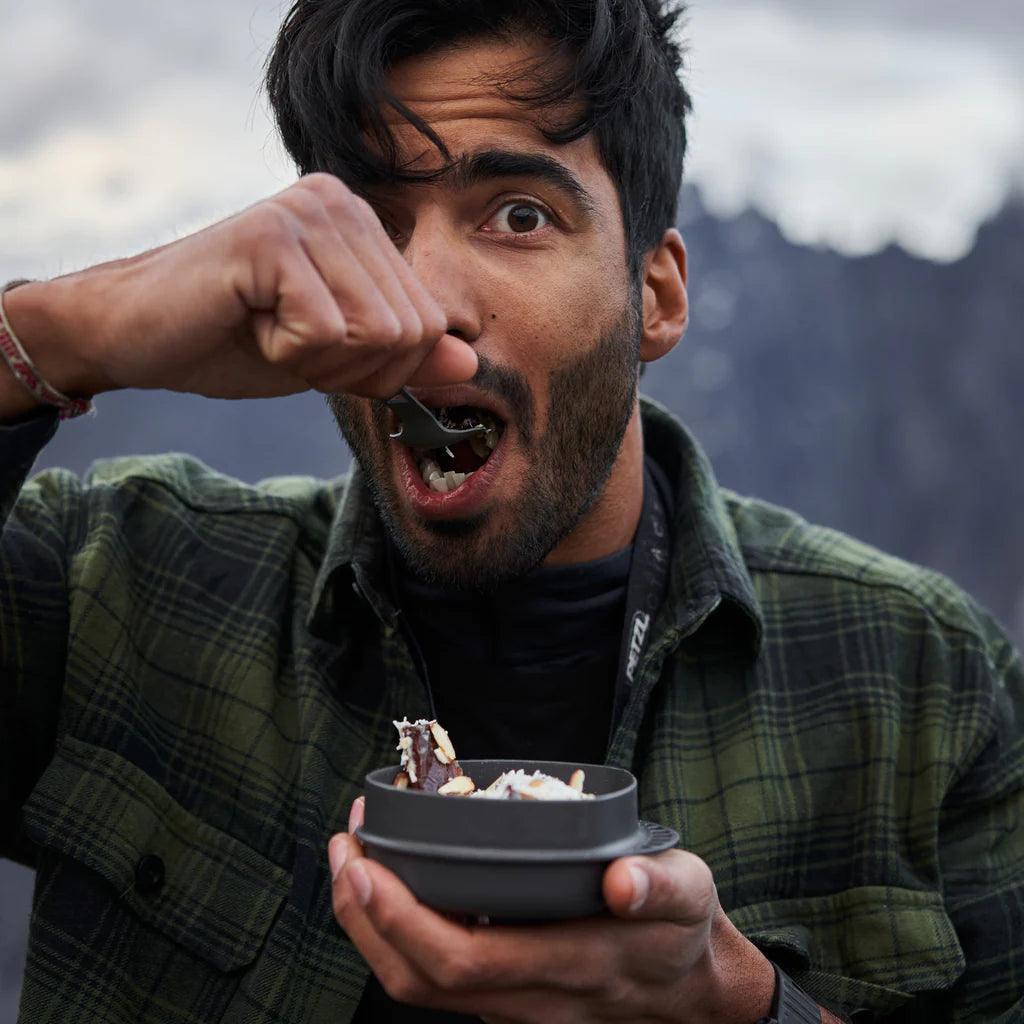 Man eating outdoors from a camping bowl with mountains in the background