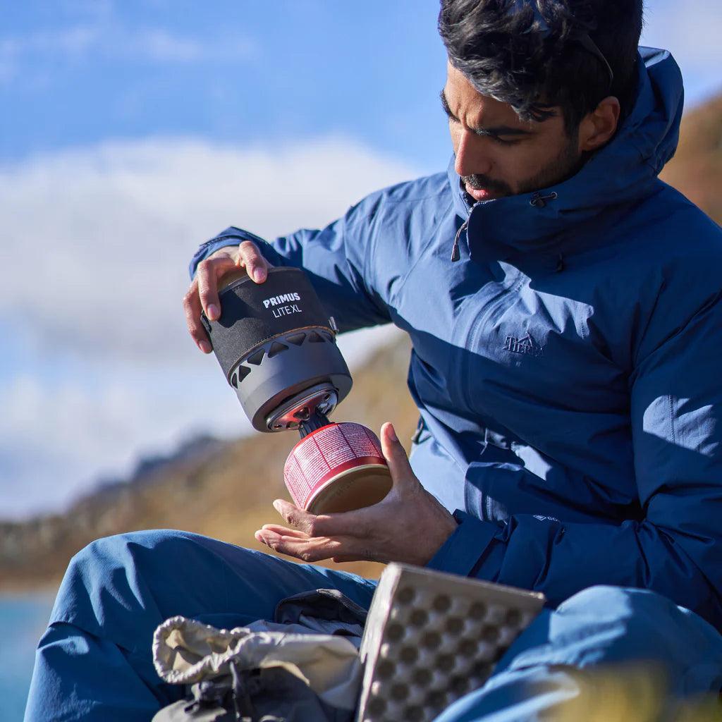 Man outdoors assembling Primus Lite XL stove with fuel canister, wearing blue jacket