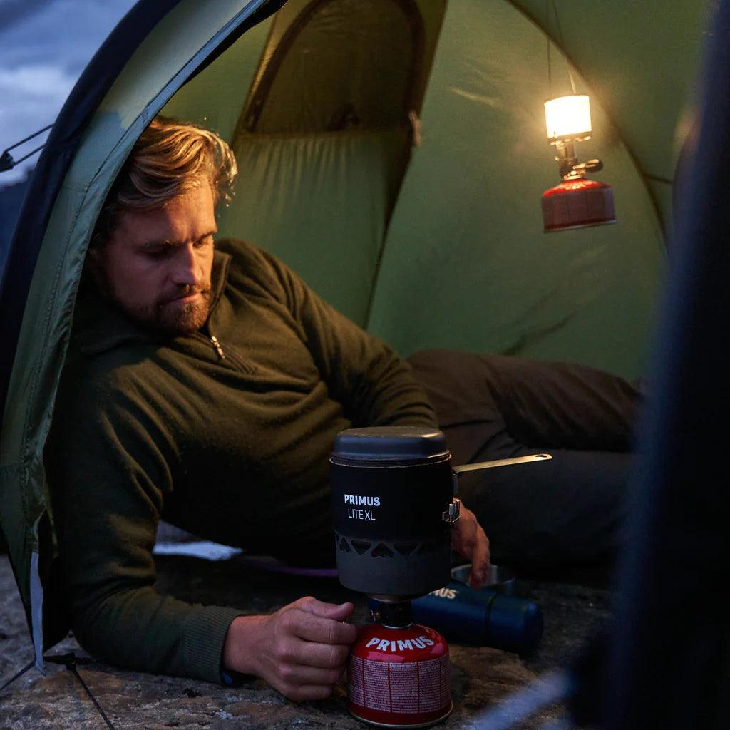 Camper using Primus Lite XL stove inside green tent with hanging lantern at dusk