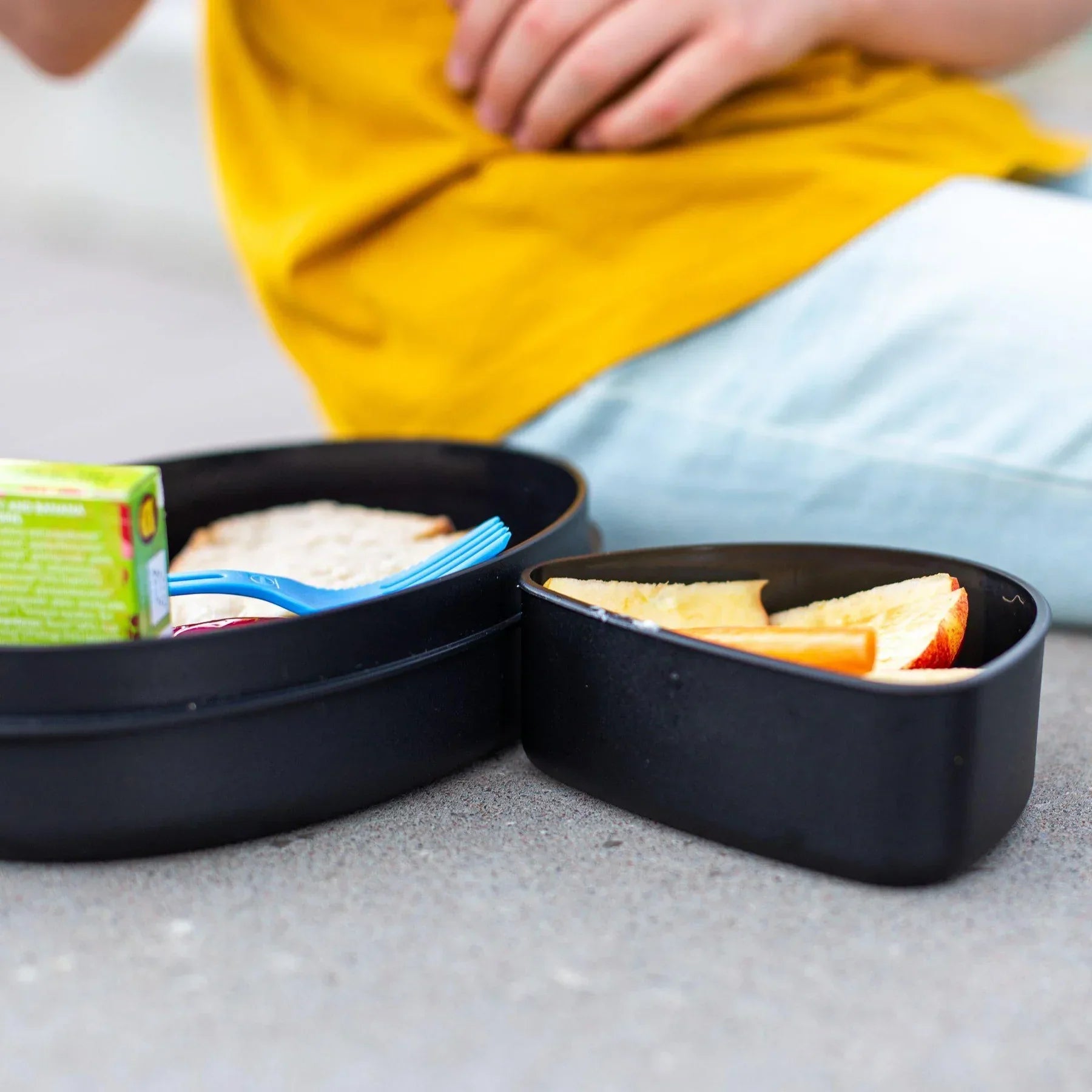 Black meal set with sandwich, juice box, fruit slices, and blue utensil outdoors