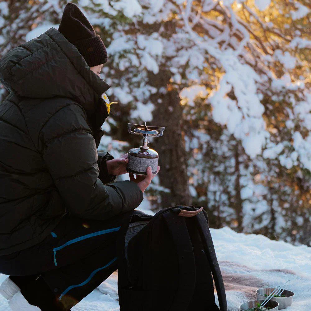 Camper holding portable gas stove outdoors in snowy forest with winter backpacking gear