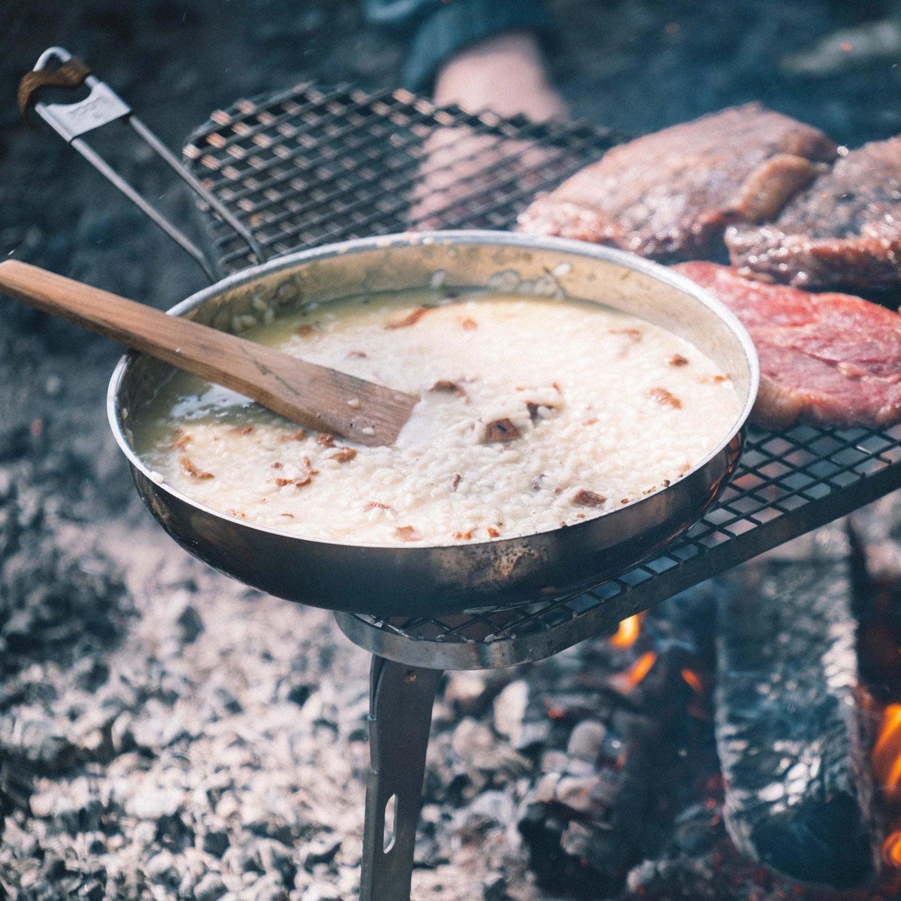 Risotto and steaks cooking on a metal grill over an open fire outdoors