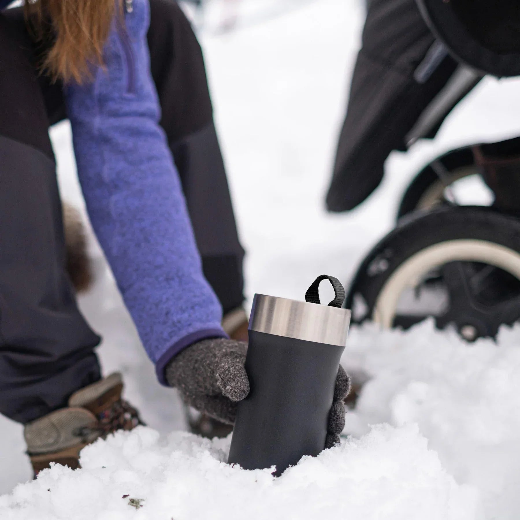 Person in winter clothing holding black stainless steel travel mug in snow near stroller