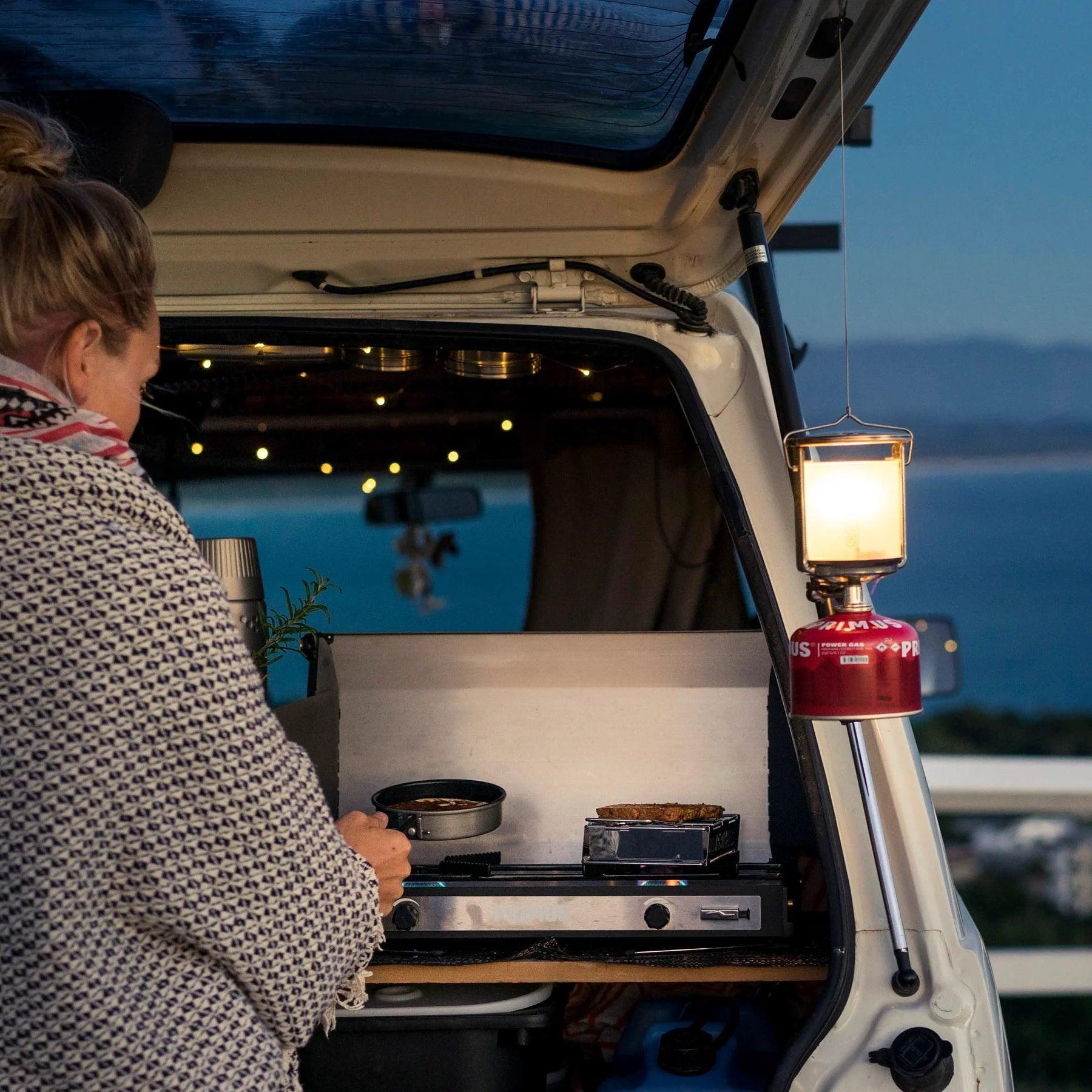 Person cooking on a portable Primus toaster in a van at a coastal campsite