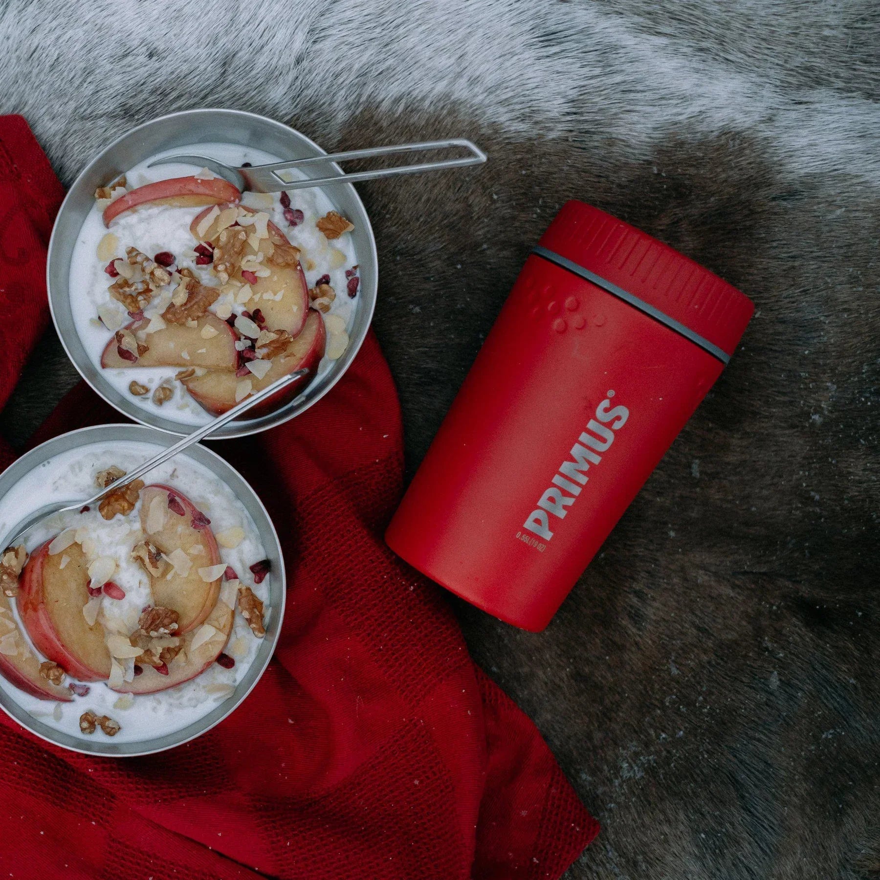 Red Primus lunch jug with two bowls of yogurt, fruit, and nuts on red fabric outdoors