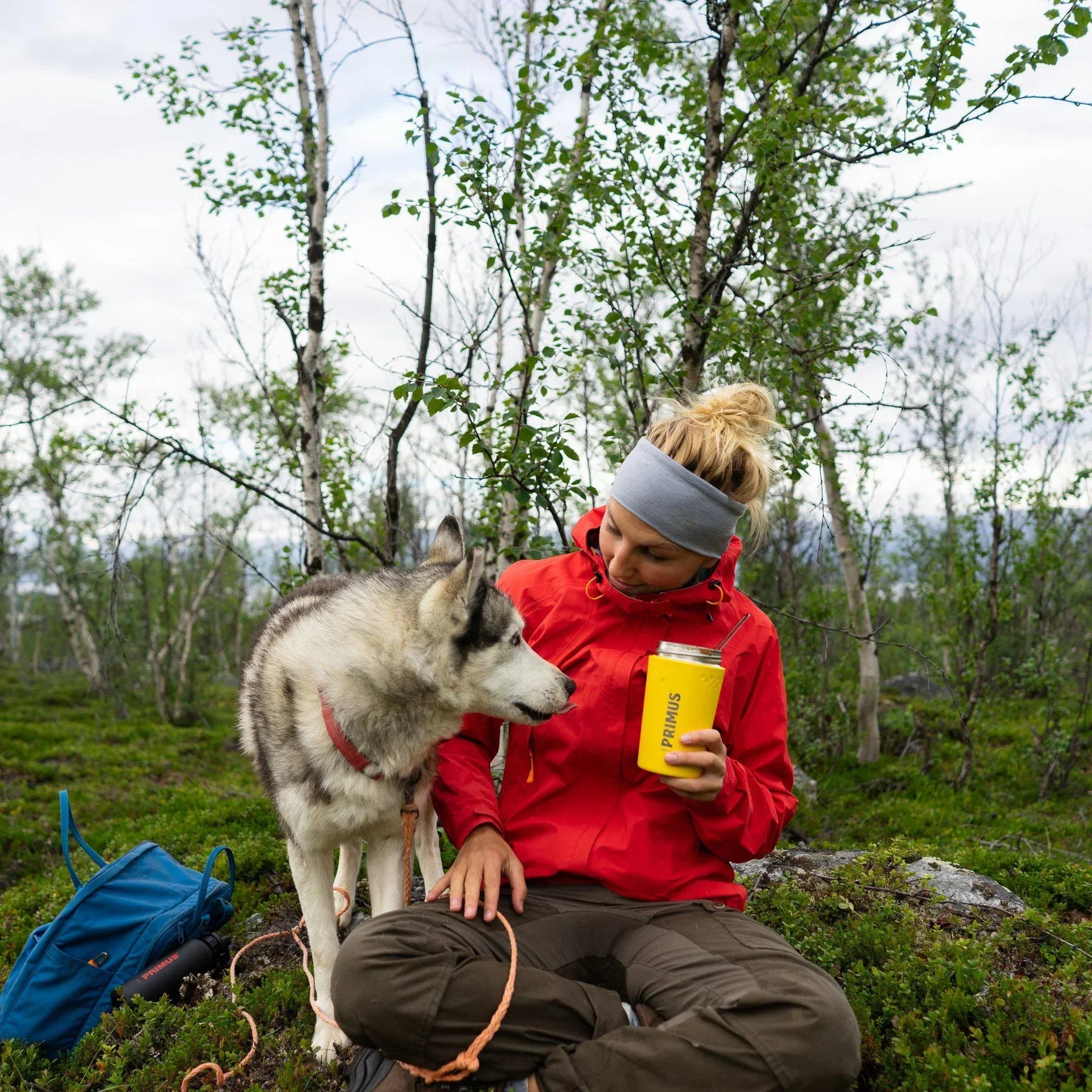 Woman in red jacket holding yellow Primus lunch jug with husky dog outdoors in forest