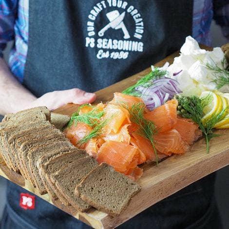 Person holding wooden board with rye bread, smoked salmon, red onions, lemon, dill, cream cheese