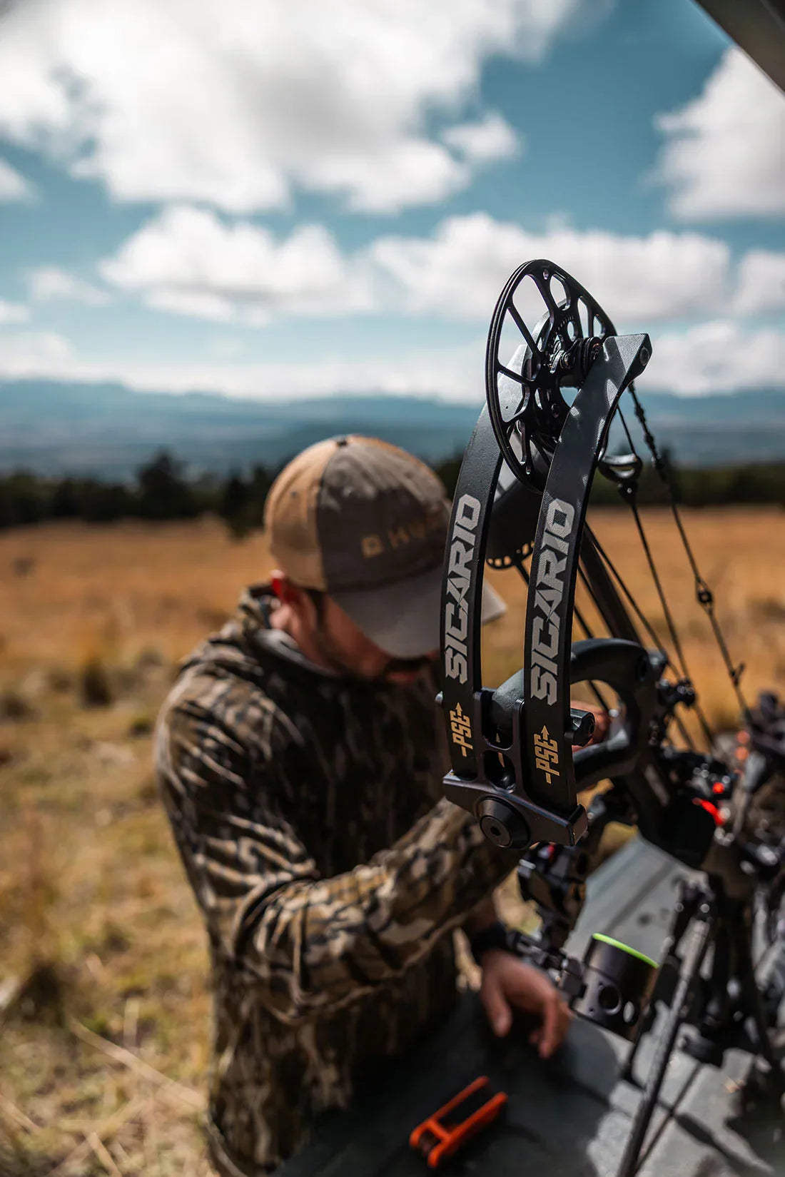 Hunter preparing PSE Sicario compound bow outdoors in camo gear with mountain view
