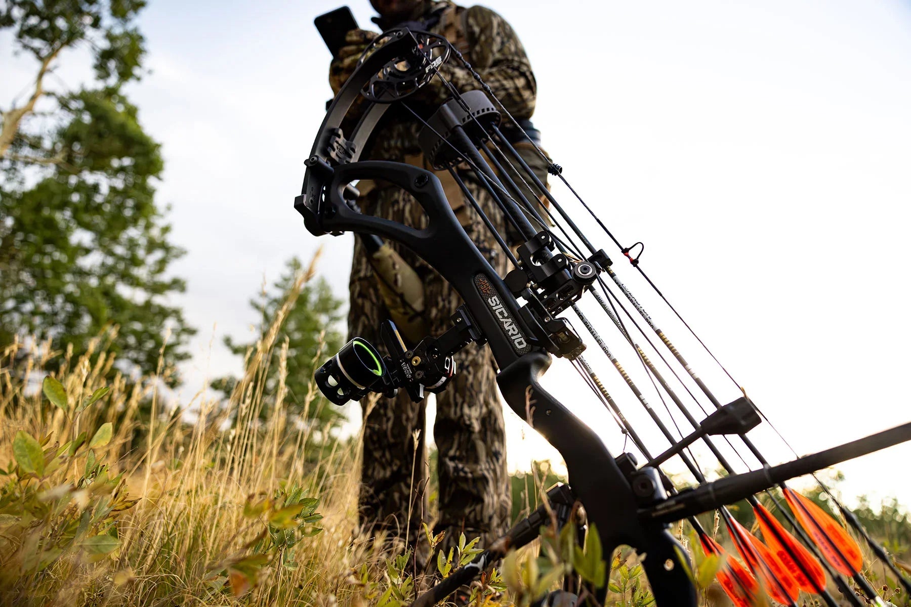 Hunter in camouflage with Sicario compound bow outdoors among tall grass and trees