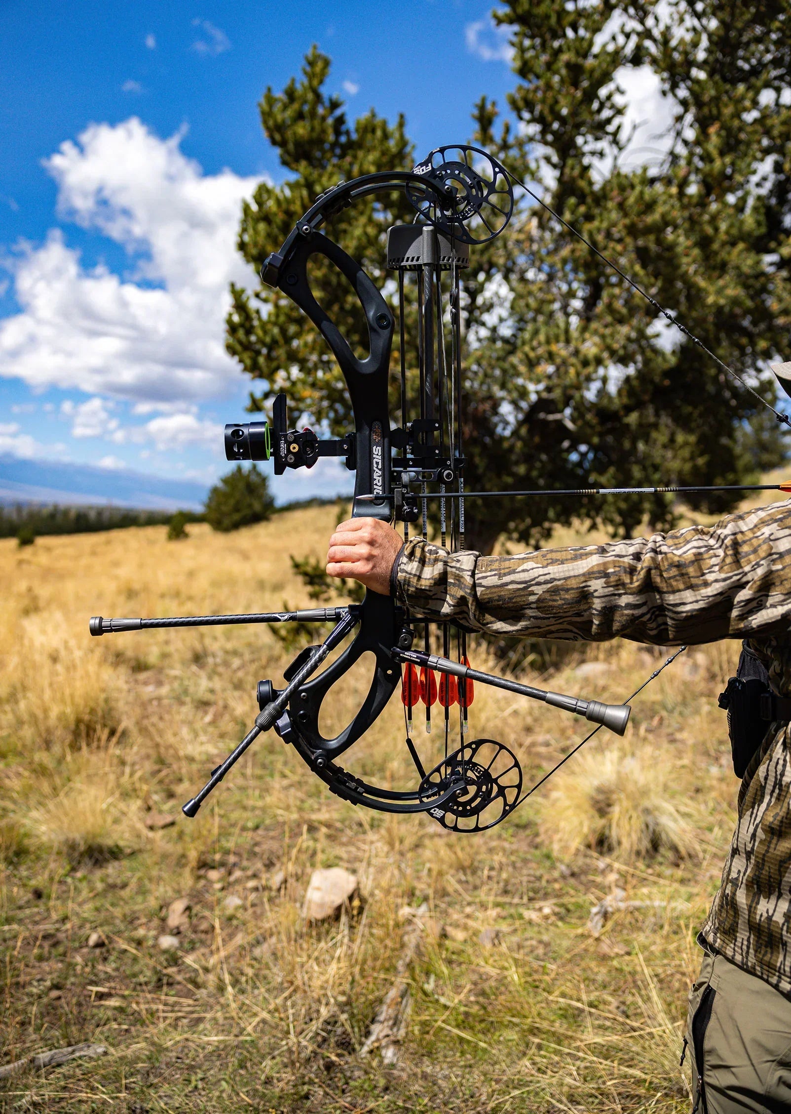 Hunter holding compound bow outdoors in grassy field with trees and blue sky