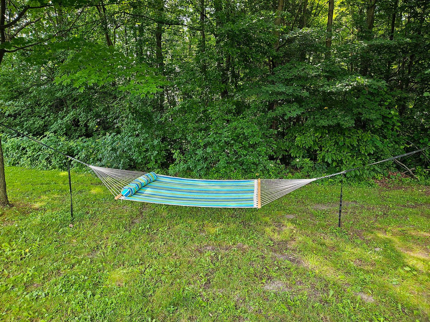 Striped blue and green hammock with pillow set up between trees in a grassy backyard