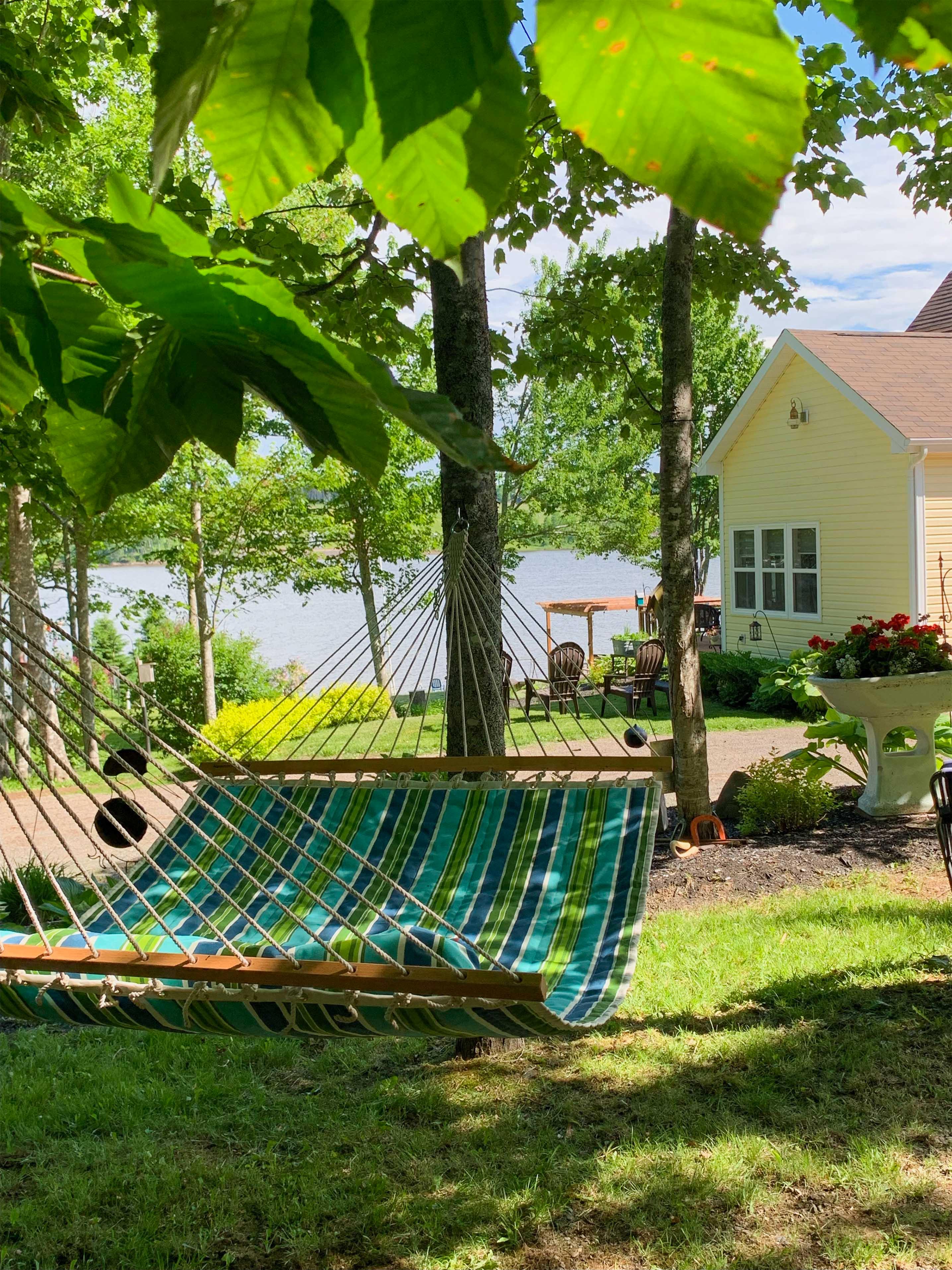 Striped hammock under leafy trees near a yellow house and lake on a sunny day