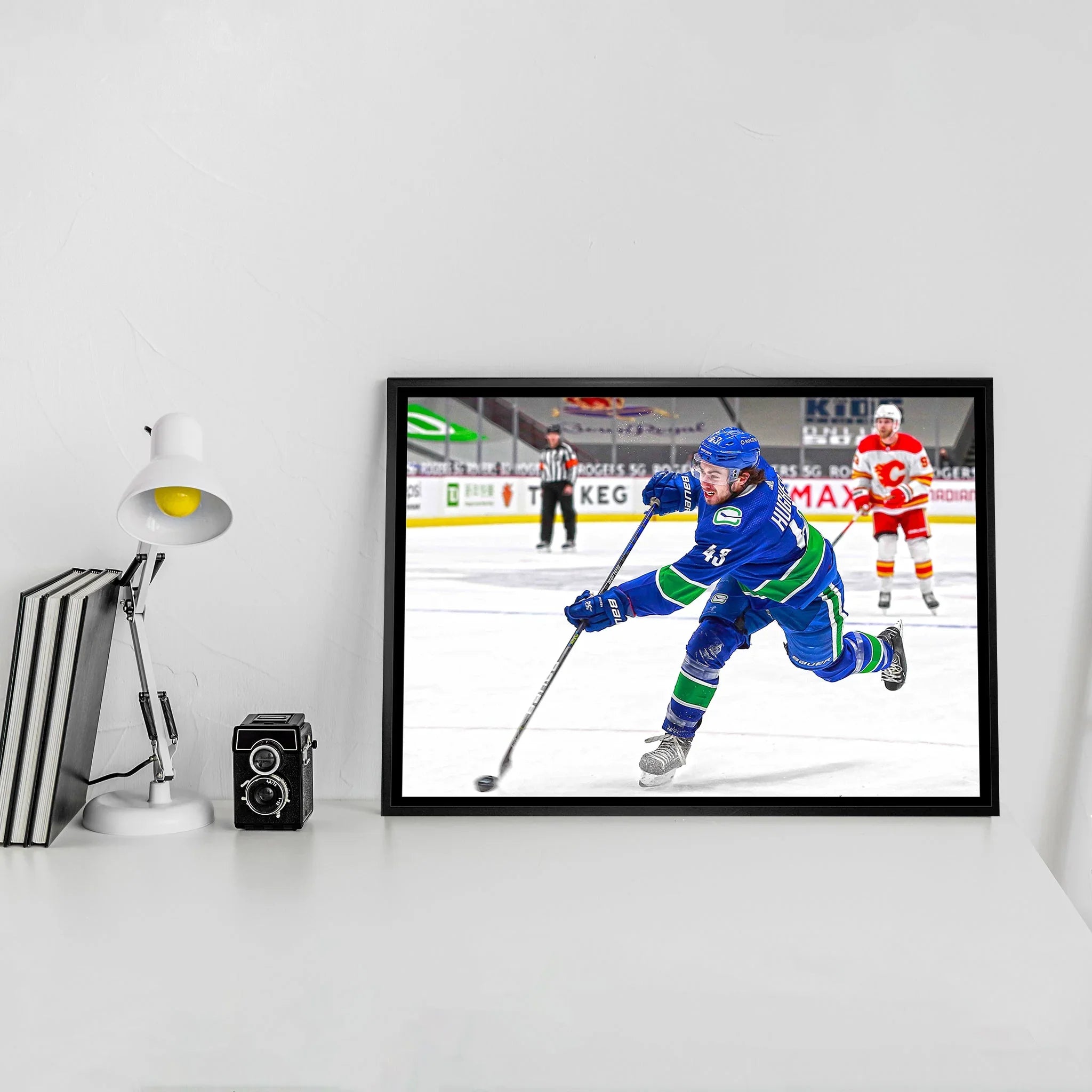 Framed hockey action photo on desk with books, camera, lamp, white wall background