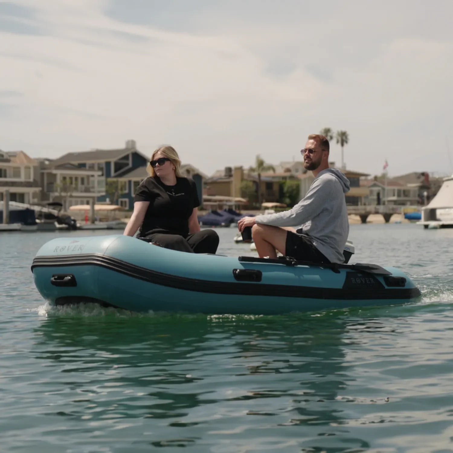 Couple riding blue Rover marine inflatable battle boat on calm water near waterfront homes