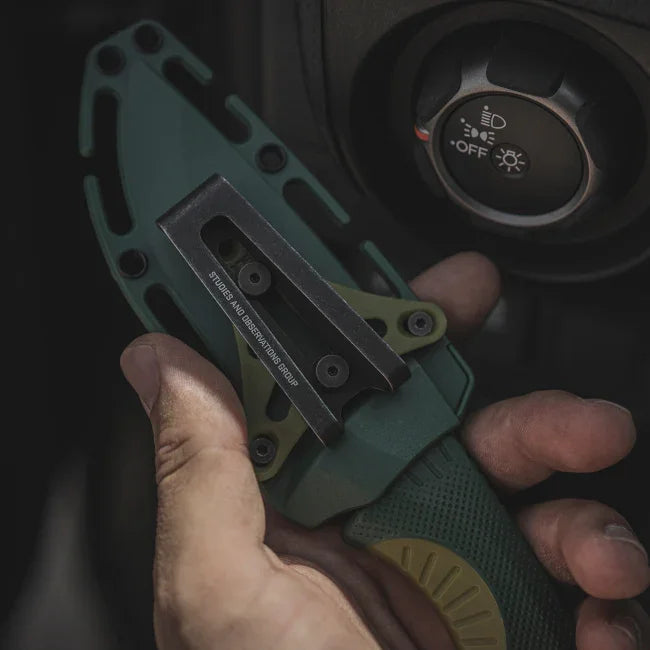 Hand holding a green tactical knife in sheath near a car headlight control knob