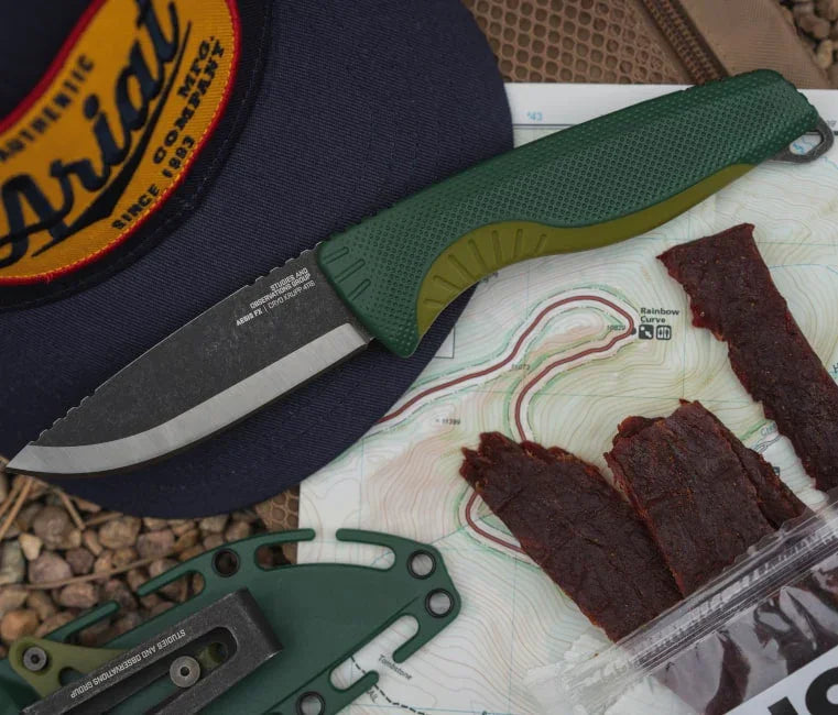 Outdoor camping knife with green handle, topographic map, and beef jerky on gravel