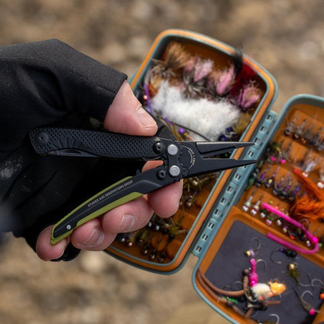 Hand holding SOG Aegis multitool pliers near open fly fishing tackle box outdoors