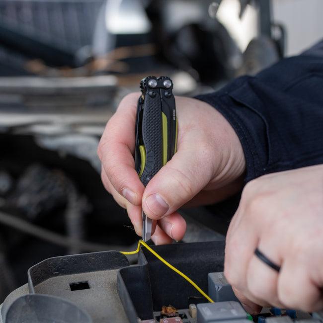 Person using a black multitool to strip yellow wire in an outdoor automotive setting