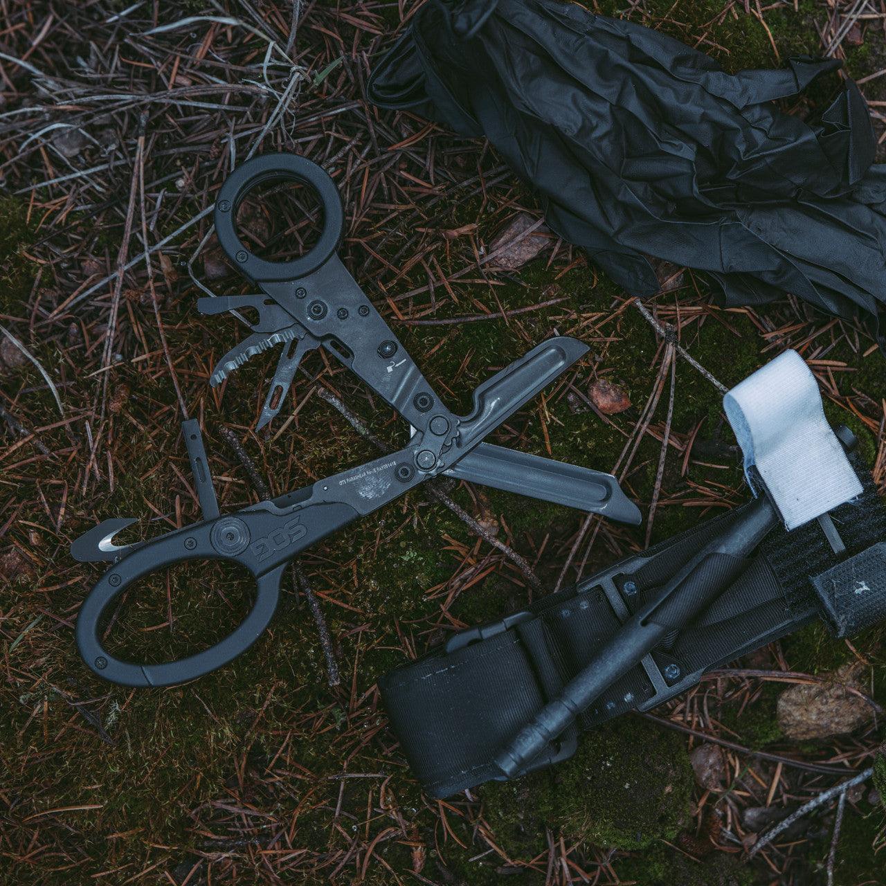 SOG Parashears multi-tool, black gloves, and sheath on forest floor outdoors
