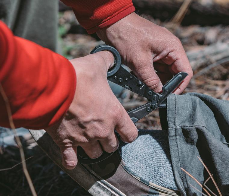 Hands using SOG Parashears multi-tool scissors outdoors to cut green fabric.