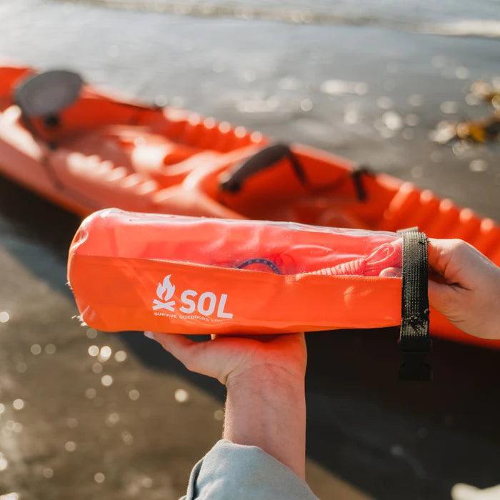 SOL survival kit held by hand near orange kayak on beach, outdoor adventure gear