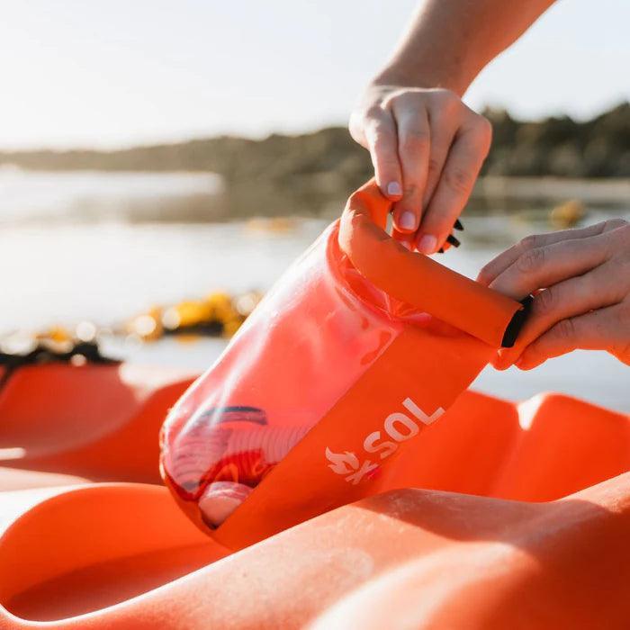 Hands opening SOL adventure survival kit bag on orange kayak outdoors by water