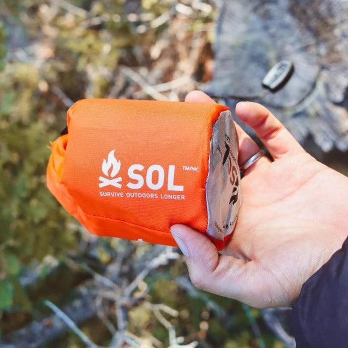 Hand holding orange SOL emergency bivvy outdoors, survival gear for camping and hiking