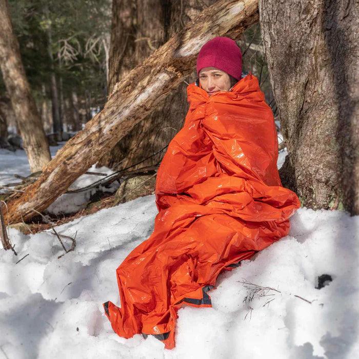 Person wrapped in orange emergency bivvy sitting in snow by trees, winter survival gear