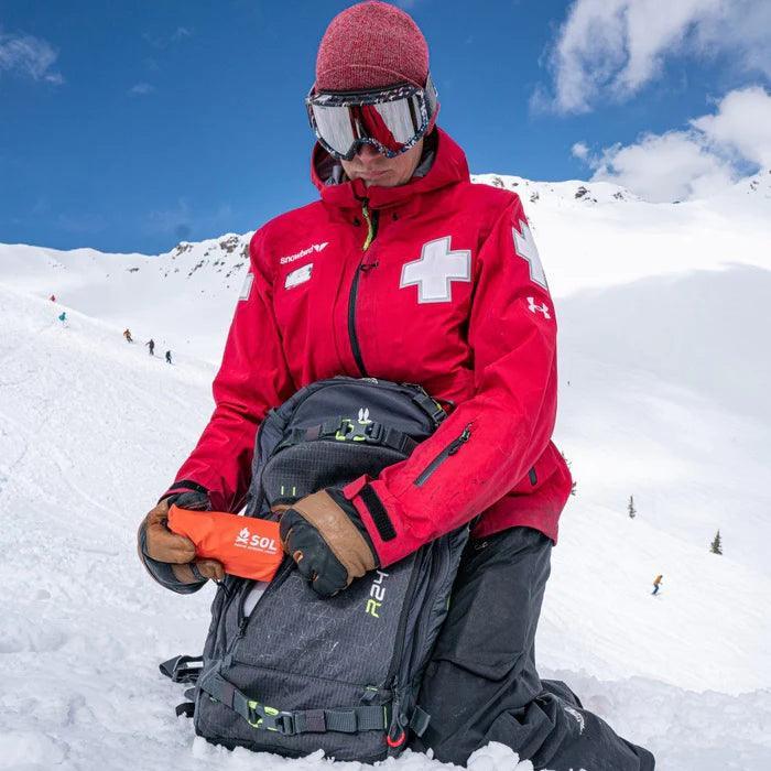 Ski patroller in red jacket with white cross holding SOL emergency bivvy on snowy mountain