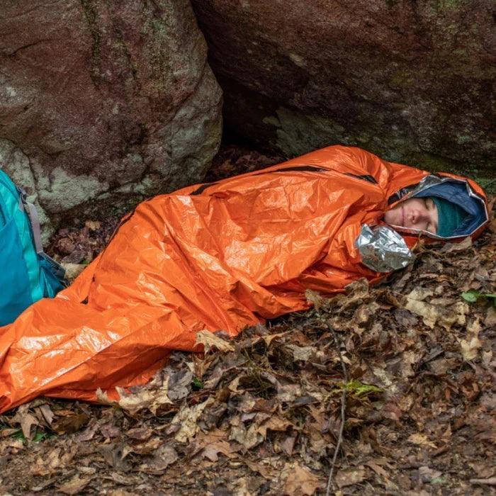 Person in orange emergency bivvy lying on forest floor near rocks, outdoor survival gear