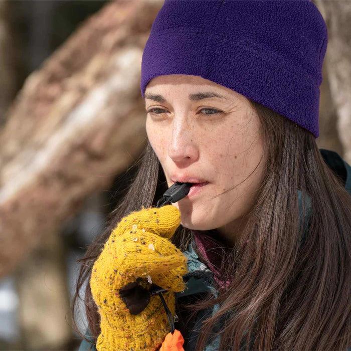 Woman outdoors wearing yellow gloves and purple beanie using emergency rescue whistle