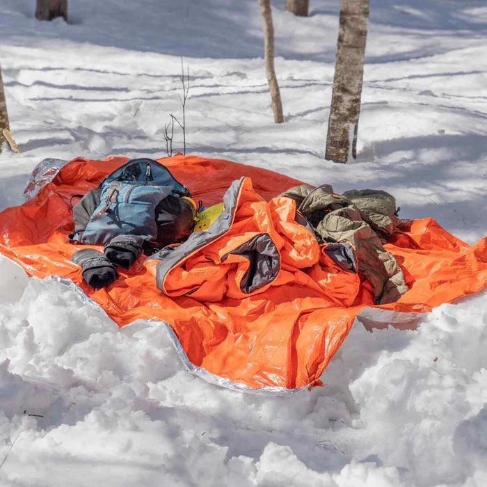 Bright orange emergency blanket and outdoor gear on snow in a winter forest setting