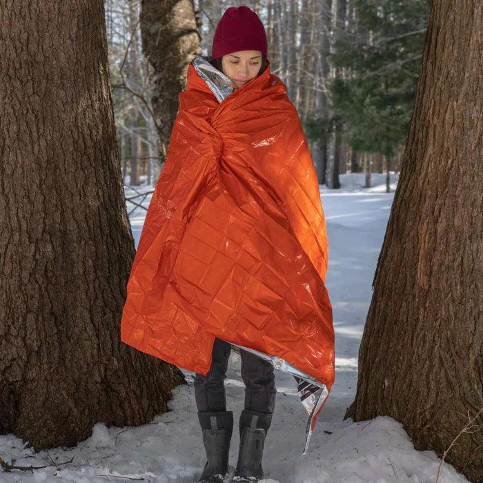 Person wearing bright orange emergency blanket outdoors in snowy winter forest