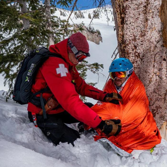 Ski patroller assists injured skier wrapped in orange emergency blanket in snowy forest