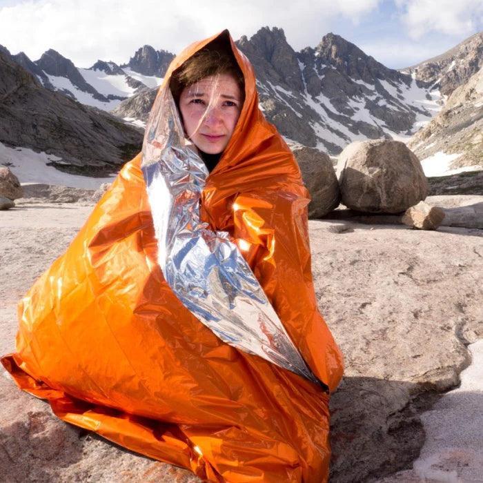 Person wrapped in orange emergency blanket outdoors on rocky mountain terrain